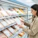 Rayon frais lumineux d’un magasin avec plusieurs emballages génériques de poulet, tandis qu’une femme compare attentivement les produits.